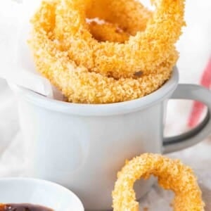 Rondelles d'oignon frites dans une tasse grise avec du papier parchemin. Il y a un petit bol en porcelaine blanche avec de la sauce barbecue à côté et une rondelle d'oignon seule se penchant contre elle.