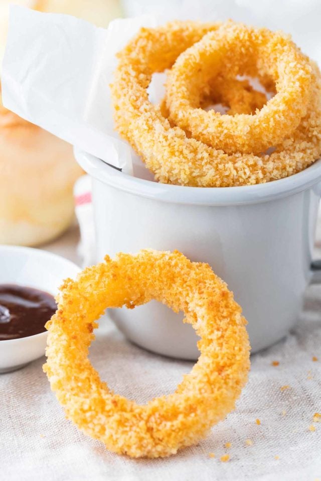 Un beignet à côté d'une tasse de café, avec des rondelles d'oignon frites dans une tasse grise avec du papier parchemin. Il y a un petit bol en porcelaine blanche avec de la sauce barbecue à côté, un oignon cru entier en arrière-plan et une rondelle d'oignon seule se penchant contre elle.
