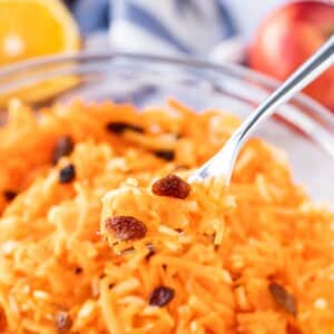 Salade de carottes avec raisins dans un bol en verre, avec une portion de celle-ci sur une fourchette surplombant le bol. Une moitié d'orange et une pomme en arrière-plan.