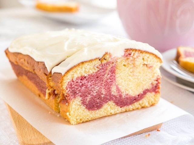 Un gâteau au yaourt aux framboises sur une planche à découper en bois recouverte de papier sulfurisé. Une tranche a été coupée et est appuyée contre. En arrière-plan, il y a une tasse rose et un empilement de deux assiettes avec une autre tranche.