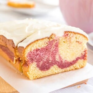 Un gâteau au yaourt aux framboises sur une planche à découper en bois recouverte de papier sulfurisé. Une tranche a été coupée et est appuyée contre. En arrière-plan, il y a une tasse rose et un empilement de deux assiettes avec une autre tranche.