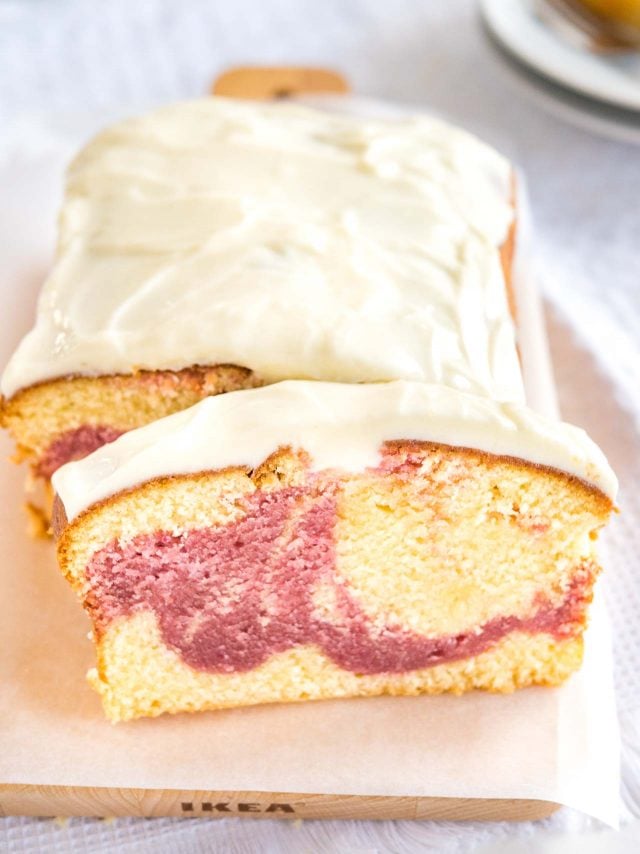 Un gâteau au yaourt aux framboises sur une planche à découper en bois recouverte de papier sulfurisé. Une tranche a été coupée et est appuyée contre. En arrière-plan, il y a deux assiettes avec une autre tranche.