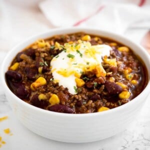 un bol blanc de chili con carne, surmonté de crème aigre, de coriandre et de fromage râpé sur une surface en marbre avec une serviette blanche et une cuillère en bronze à côté.