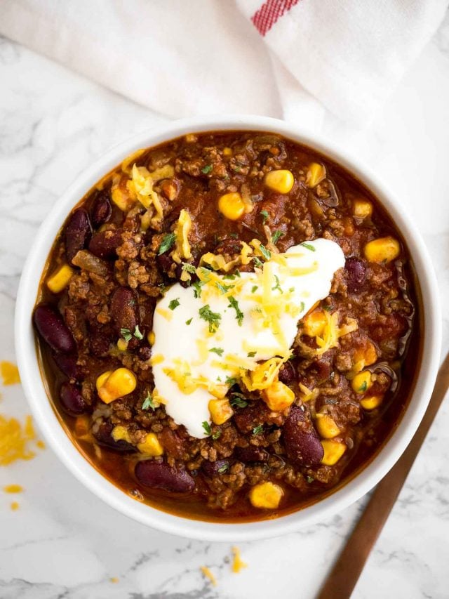 vue de dessus d'un bol blanc de chili con carne, surmonté de crème aigre, de coriandre et de fromage râpé sur une surface en marbre avec une serviette blanche et une cuillère en bronze à côté.