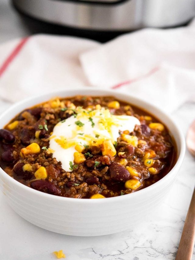 un bol blanc de chili con carne, surmonté de crème aigre, de coriandre et de fromage râpé sur une surface en marbre avec une serviette blanche et une cuillère en bronze à côté.