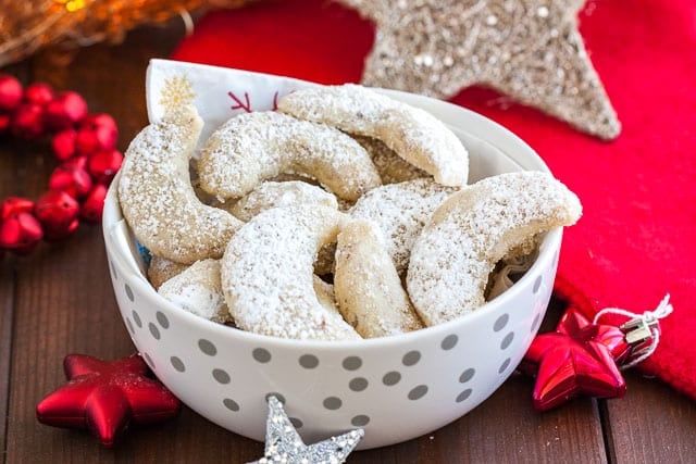 Un bol blanc avec des points argentés et une serviette, contenant des biscuits Vanillekipferl. À côté, il y a plusieurs étoiles de Noël.
