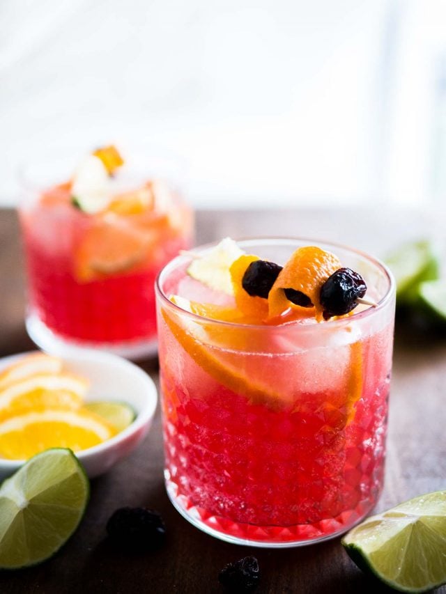 Gros plan sur deux verres de Moscow mule aux oranges et canneberges avec des brochettes de fruits. Il y a un petit bol blanc de quartiers de citron vert et d'orange ainsi que des citrons verts coupés à côté.
