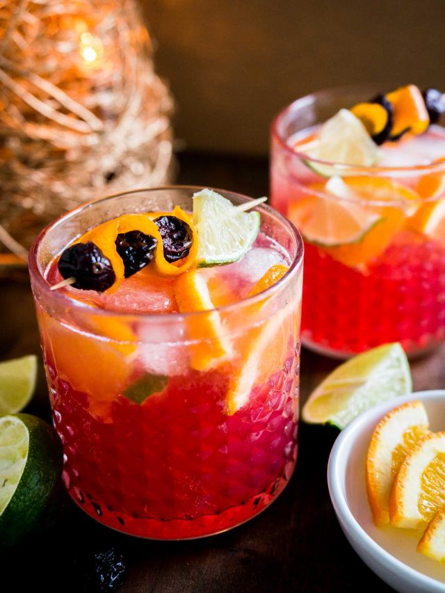 Gros plan sur deux verres de Moscow mule aux oranges et canneberges avec des brochettes de fruits. Il y a un petit bol blanc de quartiers de citron vert et d'orange ainsi que des citrons verts coupés à côté et une décoration de Noël en arrière-plan.