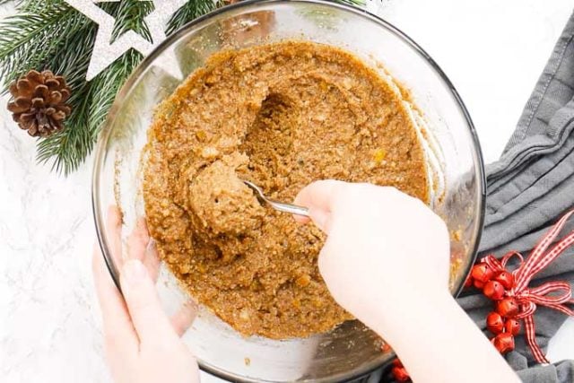 Un bol en verre avec de la pâte à lebkuchen à côté de quelques clochettes rouges et quelques branches de pin. Une main prend de la pâte avec une cuillère à café.