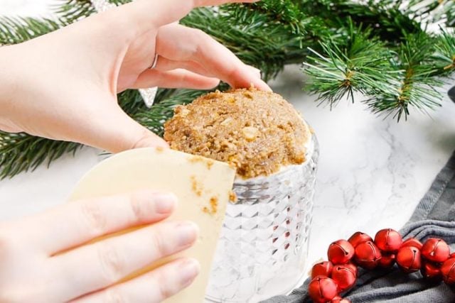 une main tenant un lebkuchen. La gaufrette est sur un verre à whisky à l'envers et la pâte est façonnée en un dôme peu profond avec une carte à pâtisserie. Il y a des clochettes rouges et des branches de pin en arrière-plan.