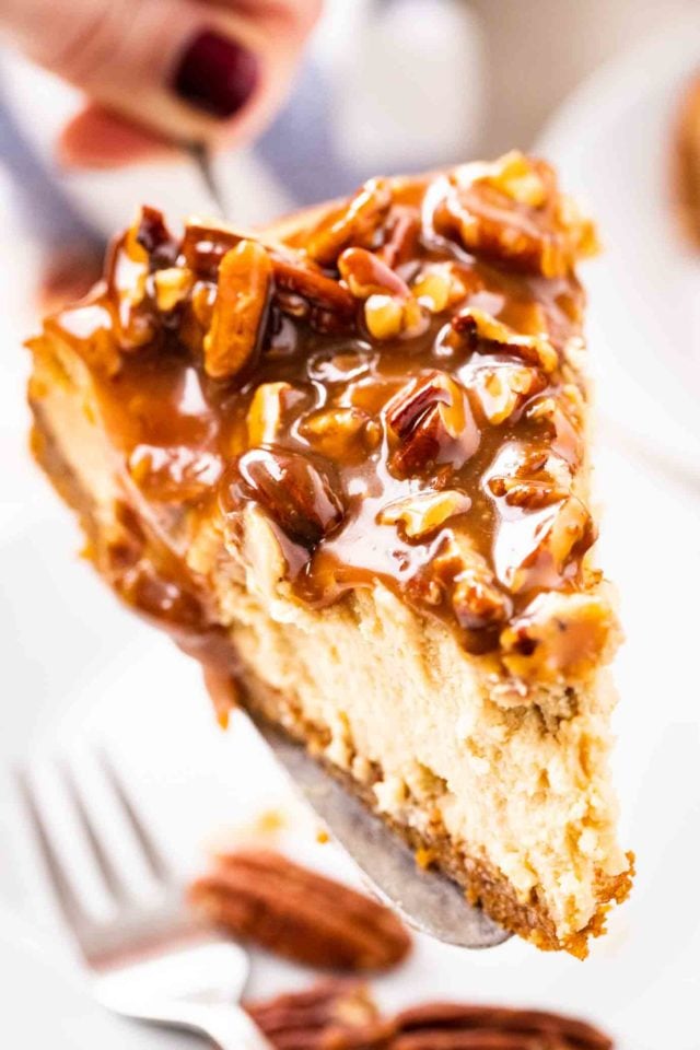 gros plan d'une part de cheesecake garnie de noix de pécan et de sauce caramel tenue sur un plat à gâteau. Il y a une fourchette et des noix de pécan entières en arrière-plan