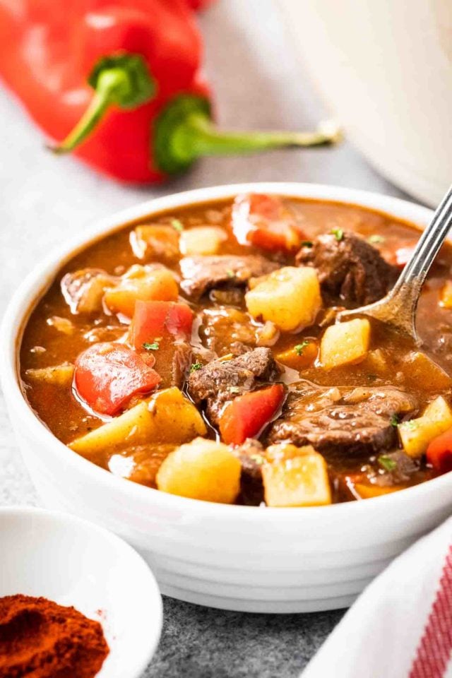 Un bol blanc de goulash, contenant des poivrons rouges, du bœuf et des pommes de terre, garni de persil et d'une cuillère. Il y a un torchon blanc avec des rayures rouges et quelques poivrons rouges à côté.