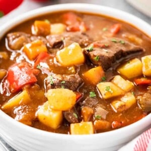 Un bol blanc de goulash, contenant des poivrons rouges, du bœuf et des pommes de terre, garni de persil. Il y a un torchon blanc avec des rayures rouges et quelques poivrons rouges à côté.