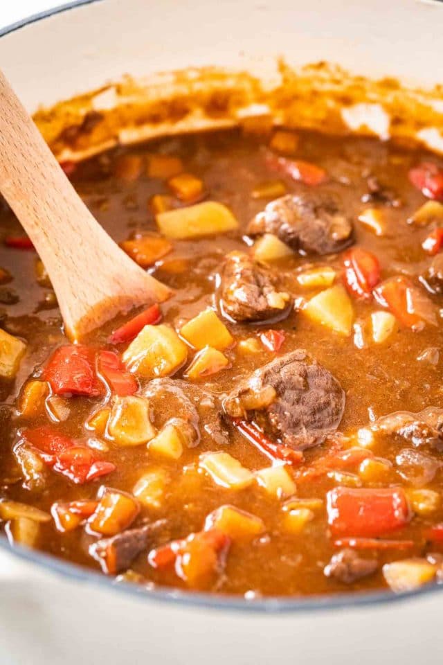 Une grande casserole avec du goulash, contenant des poivrons rouges, du bœuf et des pommes de terre avec une cuillère en bois dedans.