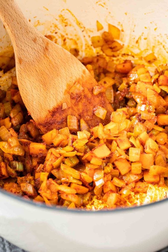 Une grande casserole avec des oignons, des carottes, du céleri et de la poudre de paprika en train de revenir avec une spatule en bois dedans.