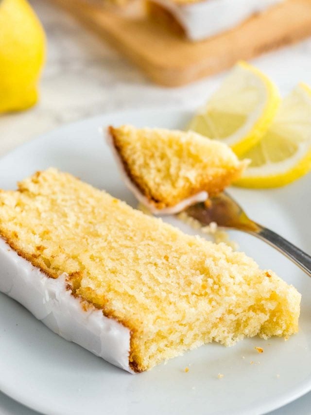 Une part de gâteau au citron moelleux avec un glaçage au citron reposant sur une assiette blanche garnie de deux quartiers de citron. Une fourchette a pris un morceau de celui-ci. En arrière-plan, une planche à découper en bois et une moitié de citron.