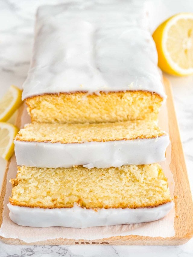 Un pain de gâteau au citron moelleux sur une planche à découper en bois, tapissé de papier parchemin. Deux tranches ont été coupées et sont posées devant. À côté de lui, il y a une moitié de citron et deux quartiers de citron.