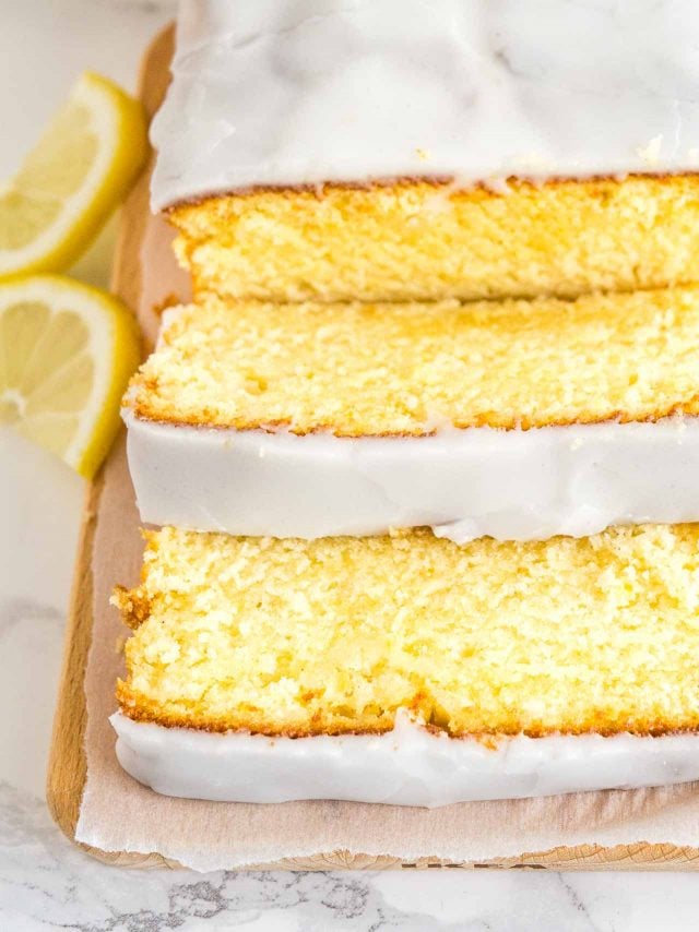 Gros plan d'un pain de gâteau au citron moelleux sur une planche à découper en bois, tapissé de papier parchemin. Deux tranches ont été coupées et sont posées devant. À côté, il y a deux quartiers de citron.