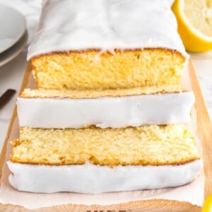 Un pain de gâteau au citron moelleux sur une planche à découper en bois, tapissé de papier parchemin. Deux tranches ont été coupées et sont posées devant. À côté, il y a une moitié de citron et deux assiettes blanches.