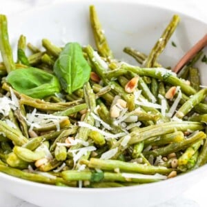 Un bol blanc de haricots verts rôtis au four surmontés de pignons de pin et de Parmigiano, garnis de feuilles de basilic.