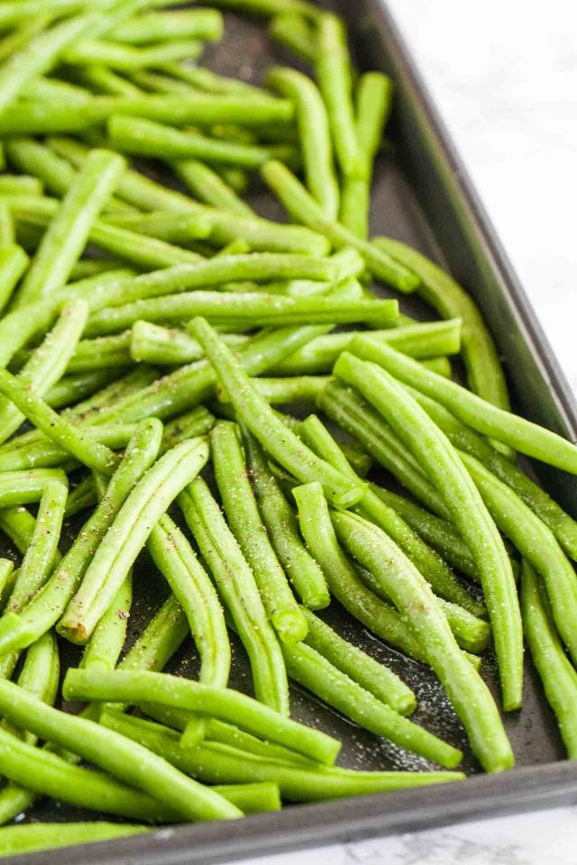 Des haricots verts non cuits sur une plaque à pâtisserie avec du sel et du poivre.