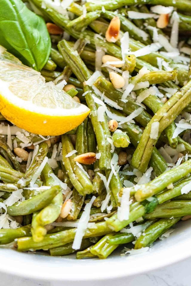 Gros plan de haricots verts rôtis au four surmontés de pignons de pin grillés et de Parmigiano, garnis d'une feuille de basilic et d'un quartier de citron.
