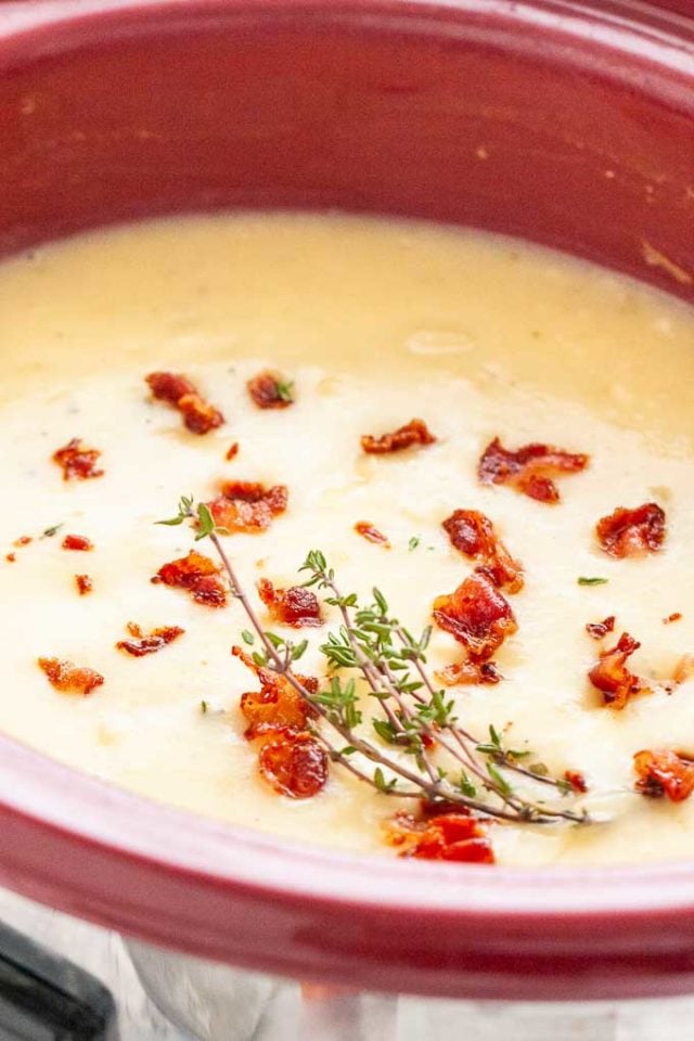 Une mijoteuse rouge remplie de soupe de pommes de terre, garnie de miettes de bacon et de thym.