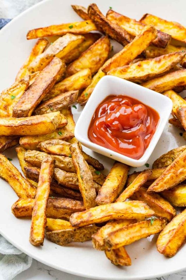 Des frites croustillantes brunes sur une assiette blanche, avec un petit bol carré blanc de ketchup.