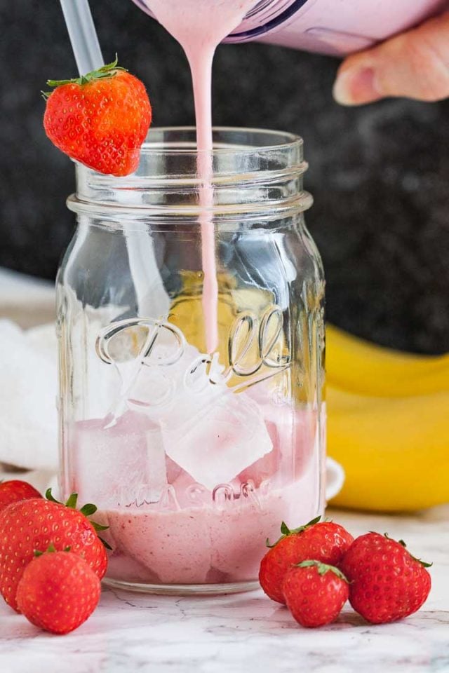 Smoothie fraise-banane versé dans un bocal avec des glaçons, une paille et une fraise sur le bord devant un fond sombre. Il y a quelques fraises et 2 bananes à côté.