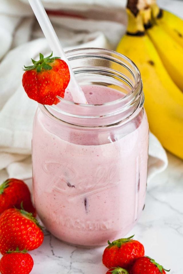 Un bocal avec un smoothie fraise-banane, une paille blanche et une fraise sur le bord sur une surface en marbre. Il y a un torchon blanc, 2 fraises et quelques bananes à côté.