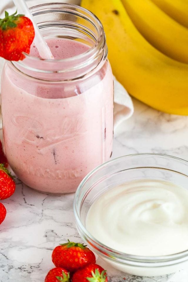 Un bocal avec un smoothie fraise-banane, une paille blanche et une fraise sur le bord sur une surface en marbre. Il y a des bananes, des fraises et un petit bol en verre de yaourt à côté.