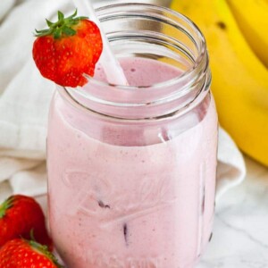 Un bocal avec un smoothie fraise-banane, une paille blanche et une fraise sur le bord sur une surface en marbre. Il y a un torchon blanc, 2 fraises et quelques bananes à côté.