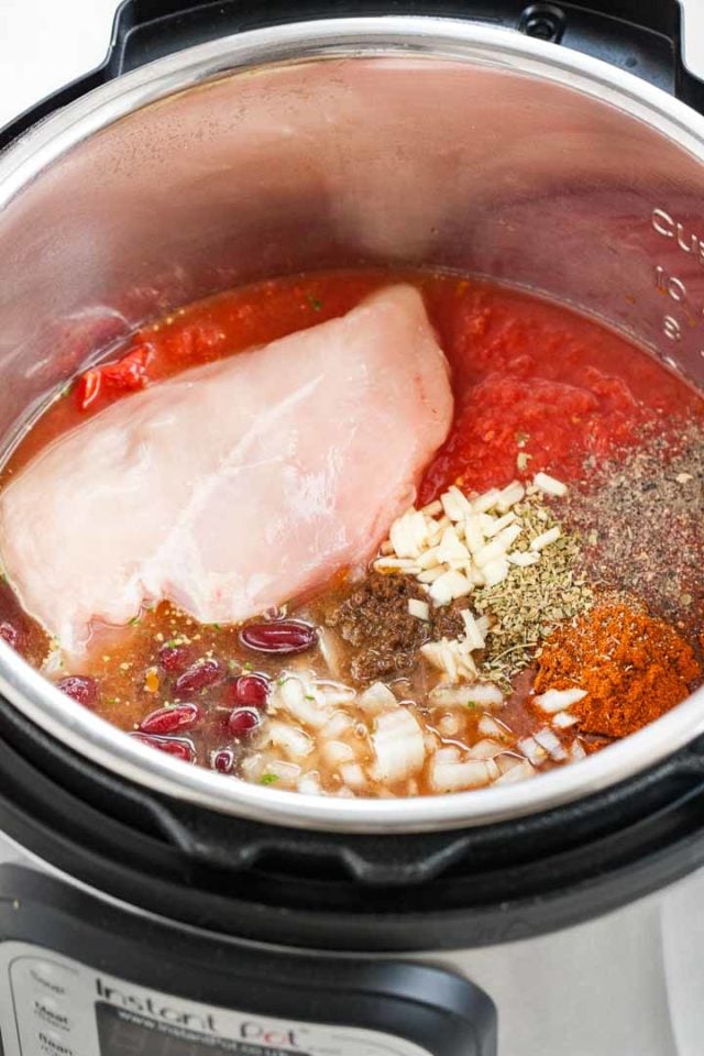 Un instant pot contenant tous les ingrédients sans les mélanger : une poitrine de poulet entière, de l'ail haché, des haricots pinto, des oignons en dés, des tomates concassées, du cumin, du poivre noir, de la poudre de chili, de l'origan.