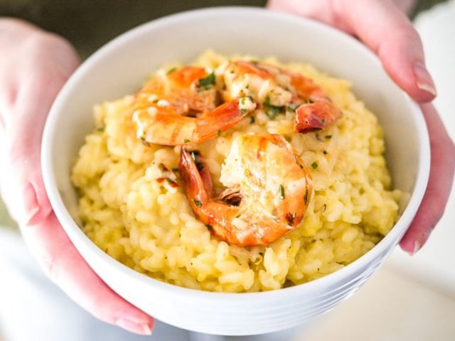 Des mains tenant un bol blanc avec du risotto au safran et des crevettes frites.
