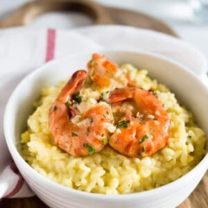 Un bol blanc avec du risotto au safran et des crevettes frites sur le dessus sur une planche à découper en bois avec un torchon blanc à rayures rouges.