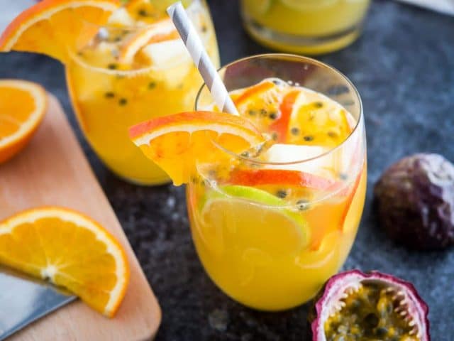 Deux verres de sangria blanche tropicale avec des tranches d'orange, des graines de fruit de la passion et une paille sur une surface sombre. Il y a des tranches d'orange, un autre verre et des fruits de la passion à côté.
