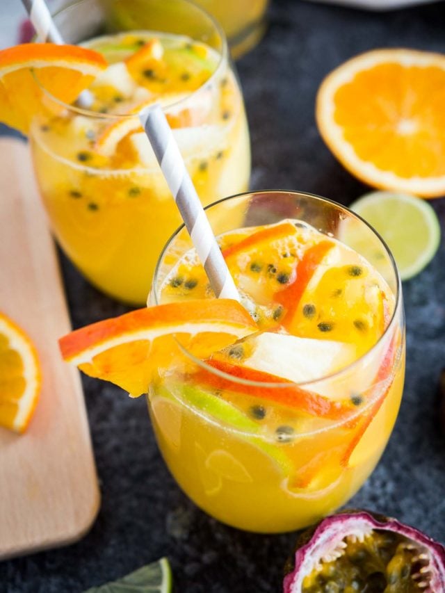 Vue de dessus d'un verre de sangria blanche tropicale avec des tranches d'orange, des graines de fruit de la passion et une paille sur une surface sombre. Il y a des tranches d'orange, un autre verre et des fruits de la passion à côté.