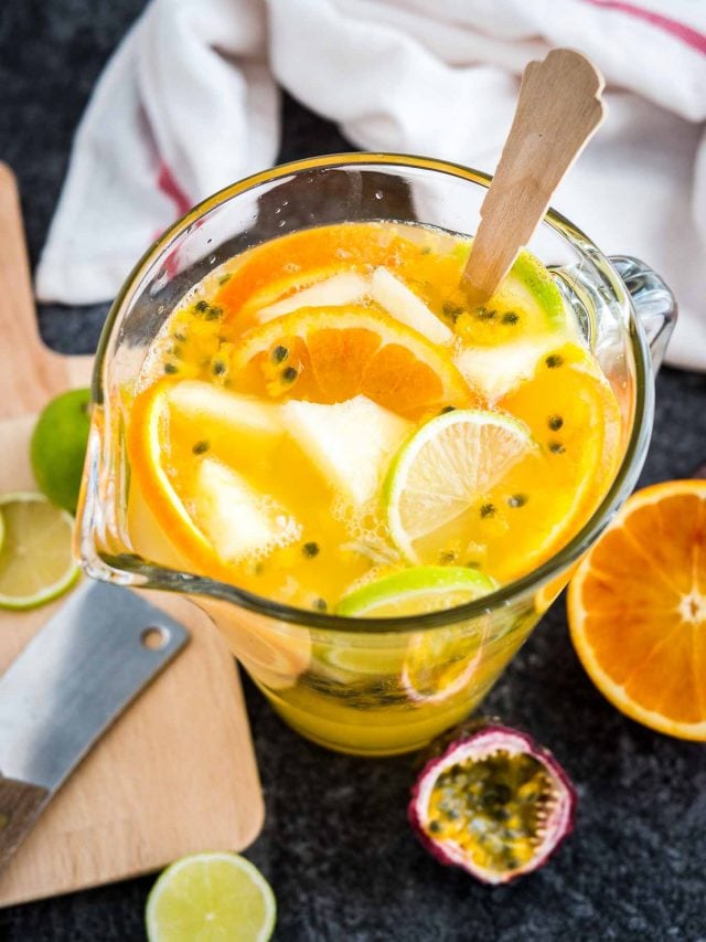 Vue de dessus d'un pichet en verre de sangria blanche tropicale avec des tranches d'orange et un fruit de la passion à côté d'une planche à découper en bois et de quelques morceaux de fruits.