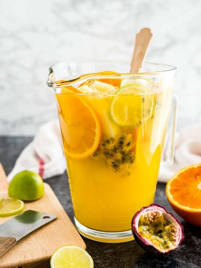 Un pichet en verre de sangria blanche tropicale avec une cuillère en bois, des tranches d'orange et un fruit de la passion. Il y a des limes, des fruits de la passion et des oranges à côté ainsi qu'une planche à découper en bois et un torchon blanc.
