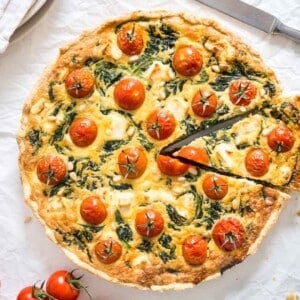 Quiche aux épinards et aux tomates sur un torchon blanc avec un couteau, un torchon rouge et blanc et quelques tomates à côté. Une part a été coupée mais est encore à sa place.