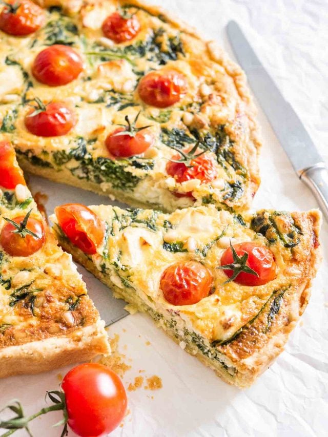 Gros plan sur la quiche aux épinards et aux tomates posée sur un torchon blanc avec un couteau et quelques tomates à côté. Une part a été coupée et tirée légèrement.