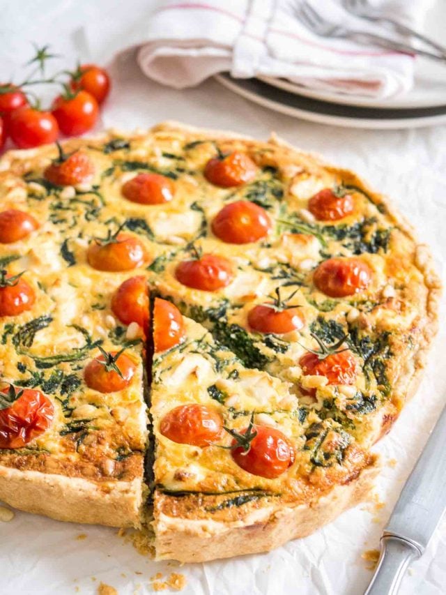 Gros plan sur la quiche aux épinards et aux tomates posée sur un torchon blanc avec un couteau, un torchon rouge et blanc et quelques tomates à côté. Une part a été coupée mais est encore à sa place.