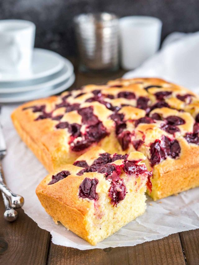 Un gâteau aux cerises avec un morceau coupé devant lui sur du papier sulfurisé. Il y a une pile d'assiettes blanches avec une tasse sur le dessus en arrière-plan.
