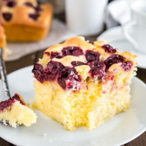 Un morceau de gâteau aux cerises sur une assiette blanche avec une bouchée prise par une cuillère. Le reste du gâteau est en arrière-plan ainsi qu'une tasse blanche.