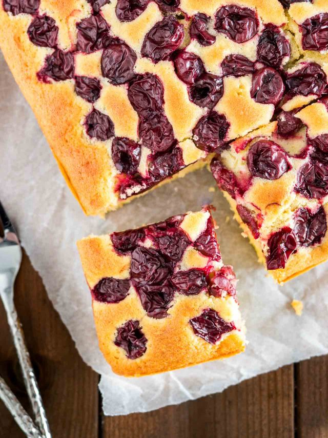 Vue de haut d'un gâteau aux cerises avec un carré coupé et placé à un angle sur du papier sulfurisé avec une fourchette.
