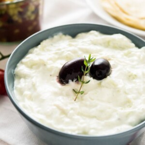 Un bol gris ardoise de Tzatziki, avec deux olives noires et un brin de thym sur un torchon blanc. Il y a une assiette blanche de tortillas et un bocal de salade grecque en arrière-plan.