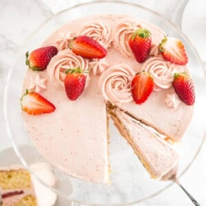 Vue de dessus d'un gâteau aux fraises sur un plat de service en verre avec une part étant soulevée par une spatule à gâteau.