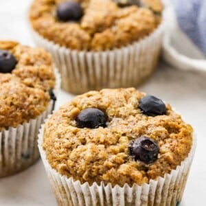 Trois muffins à la banane et aux myrtilles sur une surface en marbre à côté d'un torchon blanc et bleu.