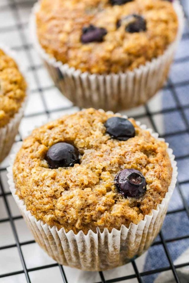 Trois muffins à la banane et aux myrtilles sur une grille de refroidissement noire sur un torchon blanc et bleu.