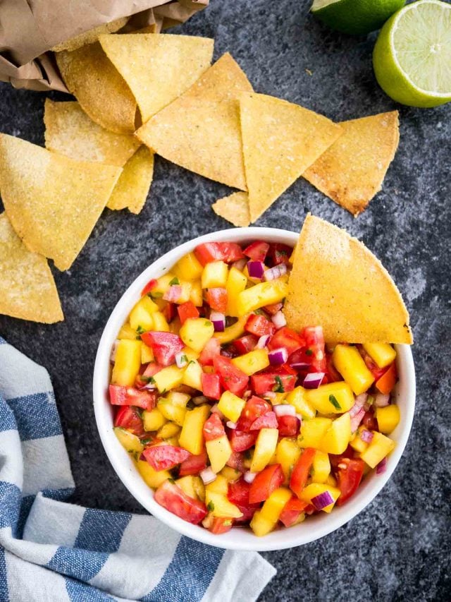 Vue de dessus d'un bol blanc contenant de la salsa aux tomates et à la mangue avec une chips à l'intérieur sur une surface sombre à côté de deux citrons verts, un sachet de nachos et un torchon blanc et bleu.